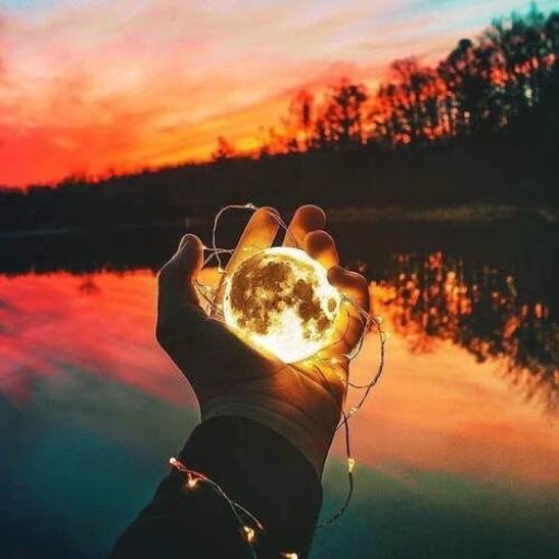 A hand holding a glowing moon-like orb by a lake at sunset