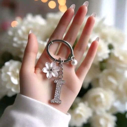 A hand holding a keychain with a letter 'I' pendant, a flower, and a pearl against a floral backdrop