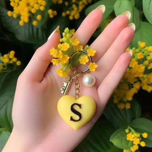 A hand holding a keychain with a yellow heart and the letter 'S', against a backdrop of small yellow flowers