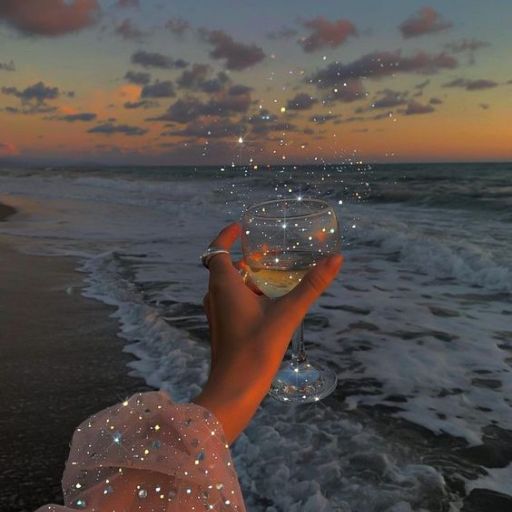 A hand holding a wine glass with glowing lights on a beach at sunset