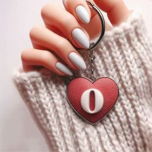 A hand with pale blue nail polish holding a keychain with a red heart-shaped charm, against a cream knitted sweater