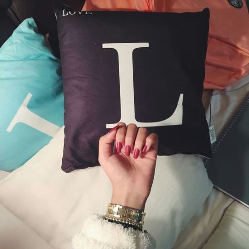 A hand with pink nails rests on a dark pillow with a white _L_ beside other colorful pillows