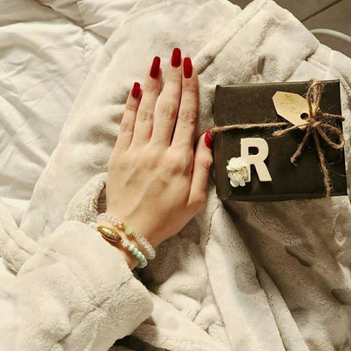 A hand with red nails on a cozy blanket, next to a black gift box with the letter 'R' and tied with twine