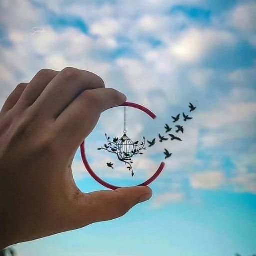 A hand holding a red ring that frames a dangling birdcage with birds flying out against a cloudy sky