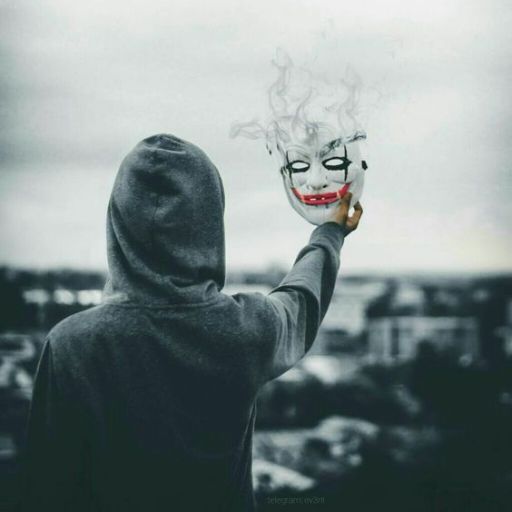 A person in a hoodie holds up a smoking clown mask against a cloudy sky