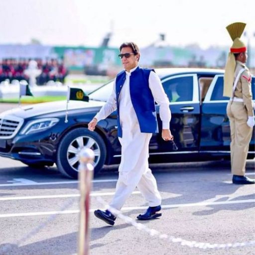 A person in traditional attire walking by a luxury car and guards on a sunny day