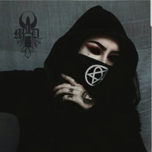 Person in a hooded black outfit with hand covering half the face, showing dark nail polish and eye makeup