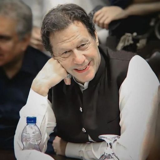 Person in white shirt and black vest sitting at a table with water bottles. Another individual in background