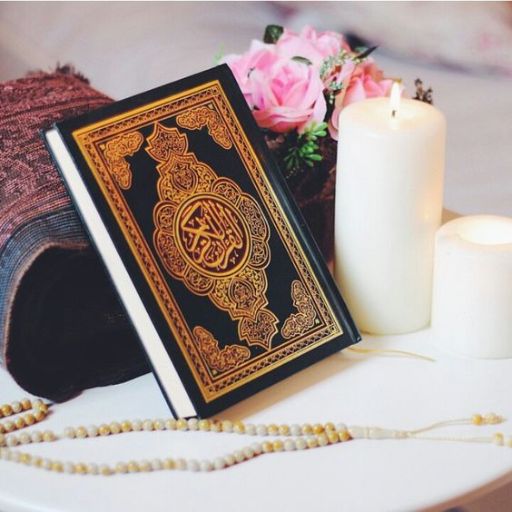 A Quran book with ornate cover beside prayer beads, candles, and a bouquet of roses on a table