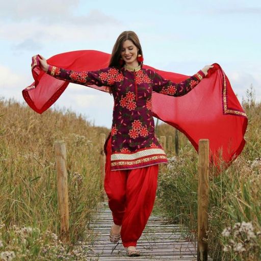 A person in vibrant red and purple traditional attire, walking on a wooden path amidst tall grasses