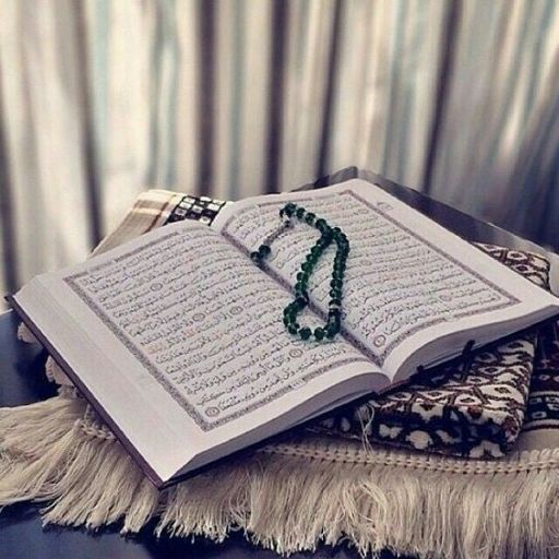 An open book, likely the Quran, with a green beaded rosary, on a patterned cloth with curtains in the background