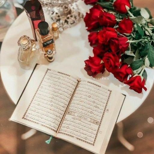 An open book, possibly the Quran, on a white table with red roses and perfume bottles nearby