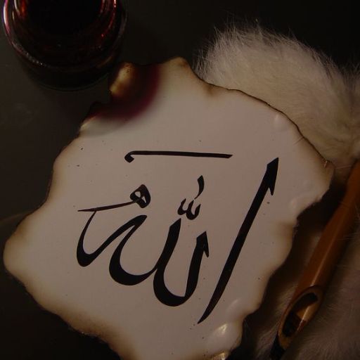 Arabic calligraphy of the word _Allah_ on a white surface with a quill and inkpot nearby